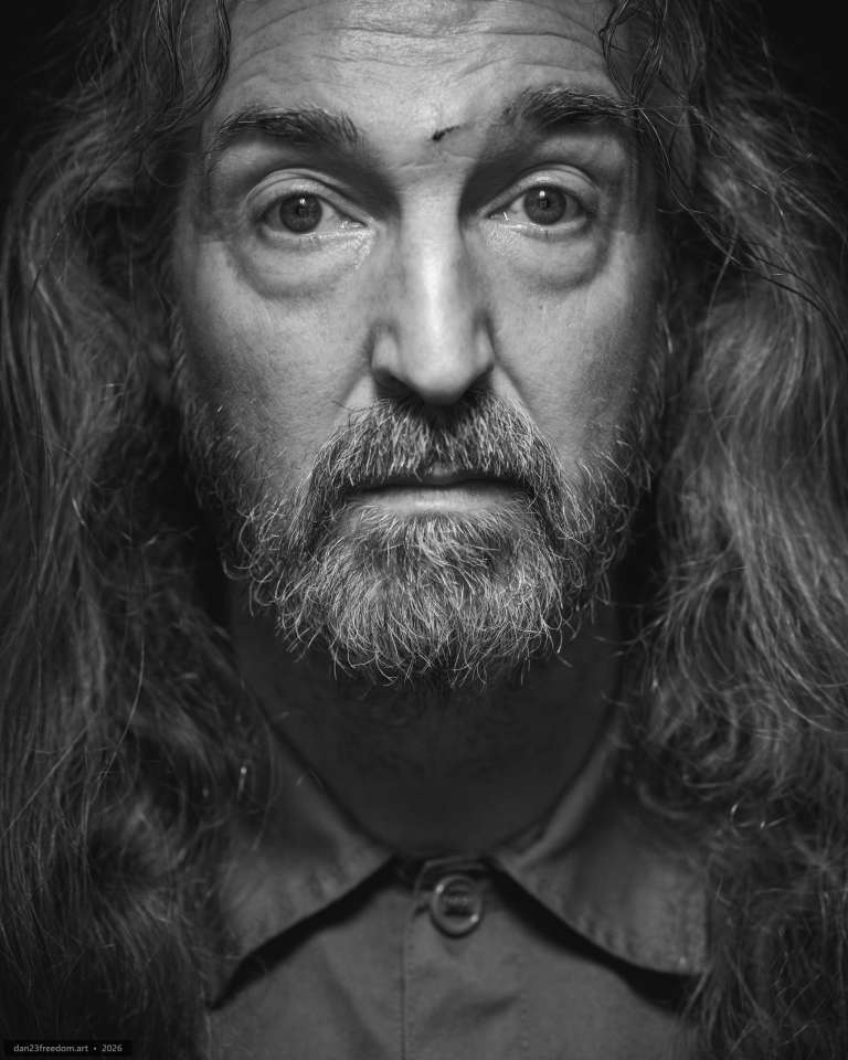 A tightly framed black-and-white self-portrait with long hair and a beard, looking directly at the camera under directional light.