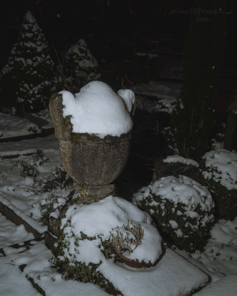 A stone urn covered in snow, sitting atop a stone pedestal in a cemetery at night. Surrounding the urn are snow-covered shrubs, a small potted plant, and ground vegetation, all blanketed in snow. The handheld flash illuminates the urn and nearby elements, creating a bright focal point against the dark, shadowy background. — Urn dreams of warm light,     Snow dances on silent stage.