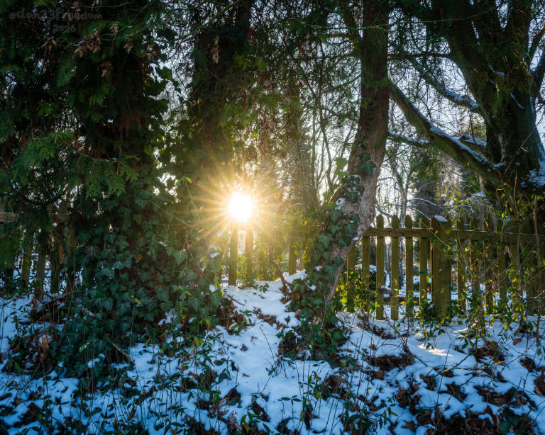 A sunburst shines through a snow‑topped picket fence at the edge of a cemetery, casting crisp rays across the winter scene.  — The sun found a gap in the fence and sent a postcard of light; the snow replied with silence.