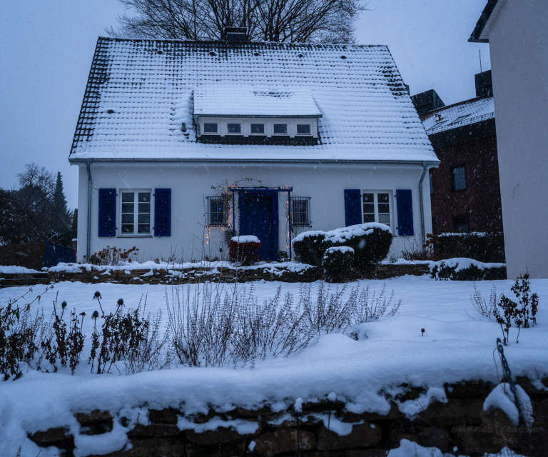 A small white house with blue shutters and string lights sits blanketed in snow, as delicate snowflakes drift down on an overcast winter day. It seems to offer a quiet, friendly greeting to those passing by.
