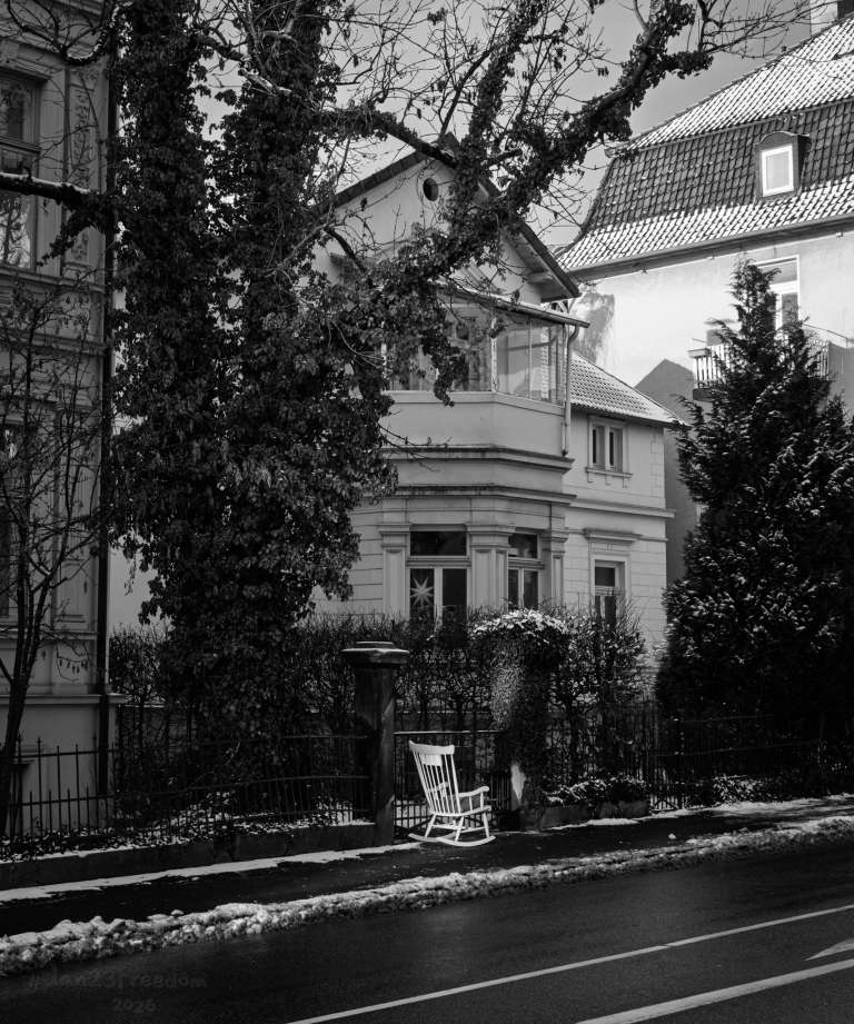 A white rocking chair sits on a snowy sidewalk outside a classical house with wrought iron fencing. The scene feels quiet and expectant, as if the chair is waiting for someone to notice it. — Once, this chair belonged to a storyteller. Now it waits for the next tale to begin.