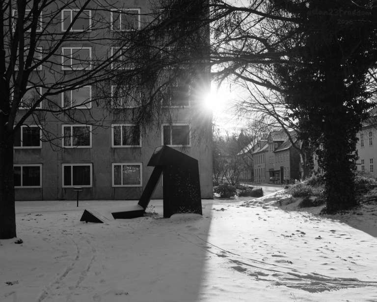 A dark metal sculpture stands in snow, partially hidden, as sunlight streams through nearby trees. The scene feels quiet and expectant, as if the sculpture is waiting for the warmth to reach it.  — The sculpture stood with snowy grace,   The sun approached, a soft embrace.