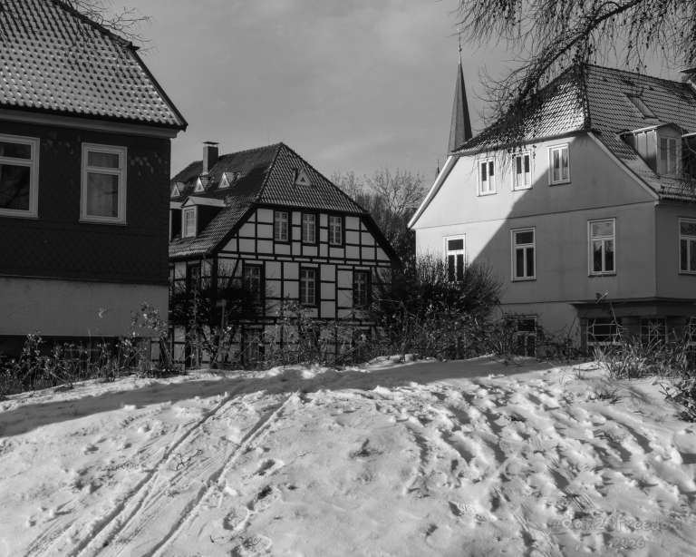 A snowy neighbourhood street with three distinct homes — the centre one half-timbered, flanked by more modern facades. Sled tracks curve across the snow, their angle echoing the shadow on the house. Footprints scatter nearby. In the distance, a church steeple rises above the rooftops. The scene feels like a pause — after play, before nightfall.