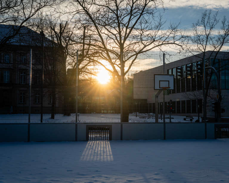 An outdoor basketball court covered in snow at sunset, with a basketball hoop and long shadows cast on the snowy surface. — Hoop waits in golden stillness, Silent court, a frozen witness.