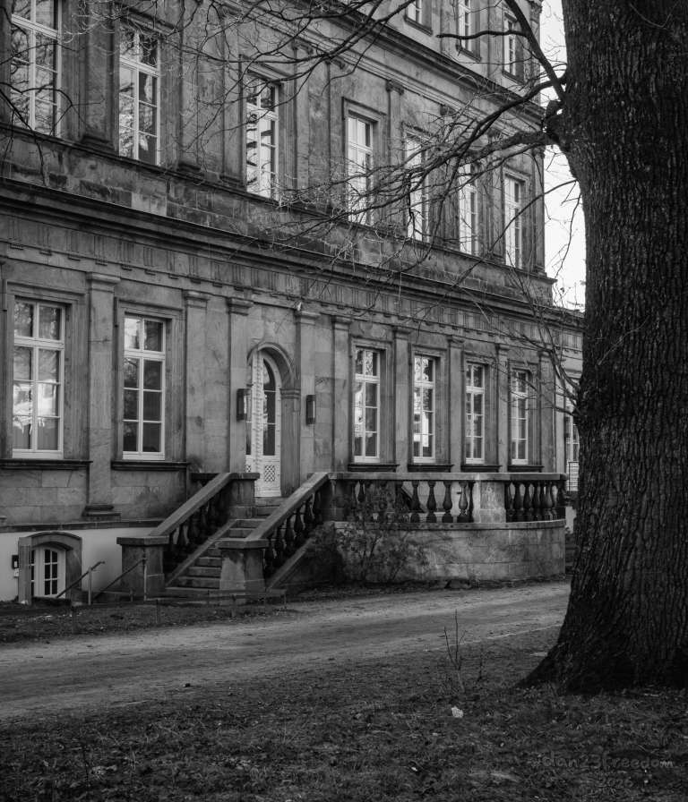 A historic stone building stands in a park, its facade catching the soft glow of late evening sun. Bare trees frame the scene, and a dirt path runs nearby. — The stone stood still, the sun leaned low,   A quiet nod before the glow.