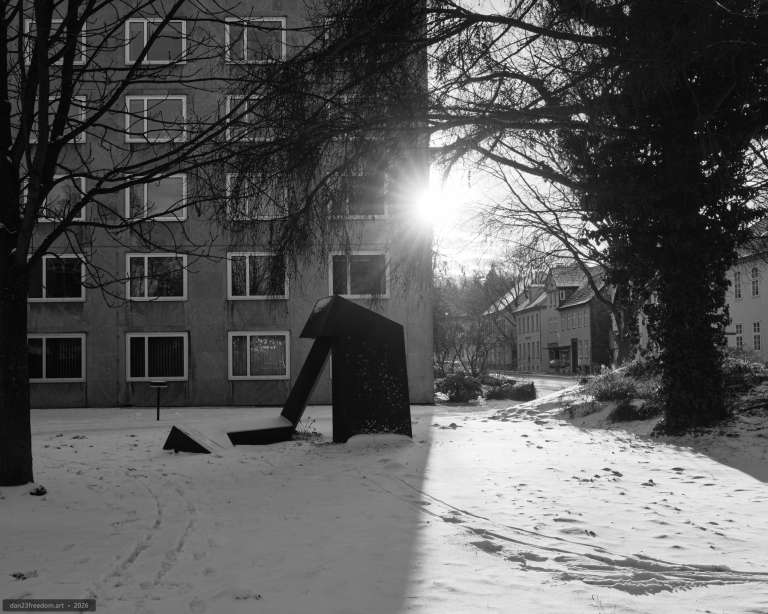 Black-and-white winter photograph of an abstract metal sculpture partly covered in snow, with footprints nearby and sunlight streaming through trees.