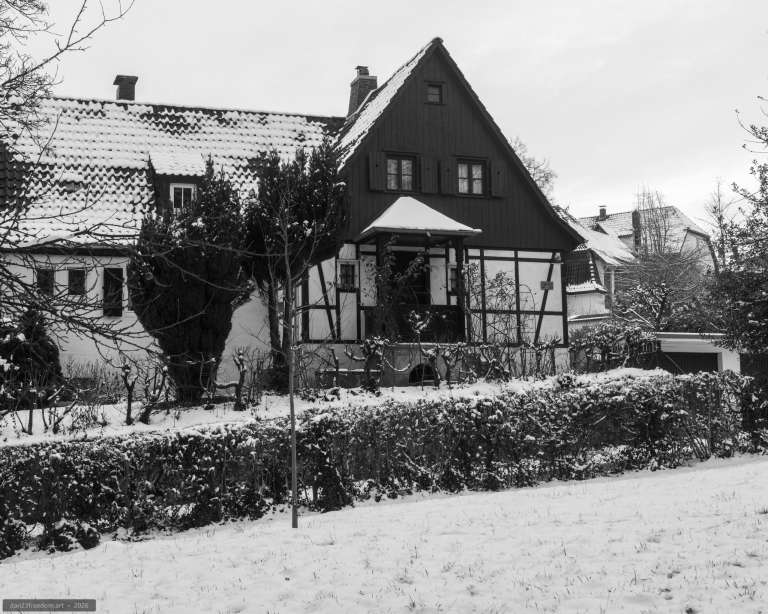 Black-and-white winter photograph of a traditional half-timbered house surrounded by snow-covered hedges, leafless trees and frost-dusted branches.