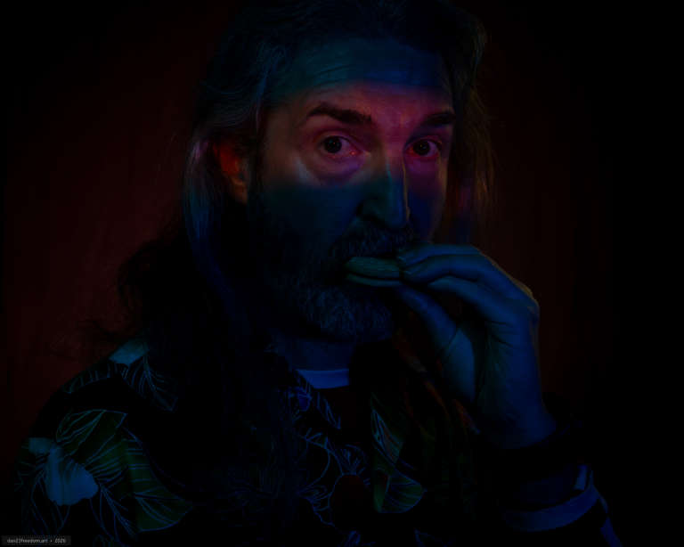 Close self-portrait against a dark background, illuminated by blue, cyan, magenta and orange light. A horizontal band of orange light crosses the eyes, and a partly eaten biscuit is held beside the mouth.